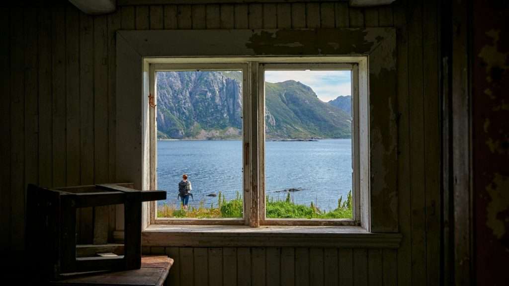 A woman stands at the water's edge, seen through an old window, pausing to take in the view of a fjord and mountains